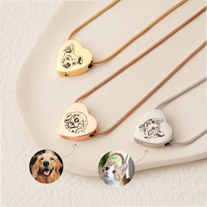 Pu&ograve; includere: Tre collane crematorie a forma di cuore con ritratti di animali domestici incisi, in oro, oro rosa e argento. Le collane sono esposte su una superficie bianca.