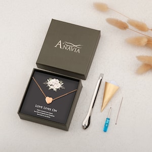 Puede incluir: Un collar con un colgante de coraz&oacute;n dorado en una caja verde con un dise&ntilde;o floral blanco. El collar est&aacute; sobre una tarjeta negra con el texto "Love Lives On. Only a moment you stayed, but what an impact your footprints have left on our hearts."