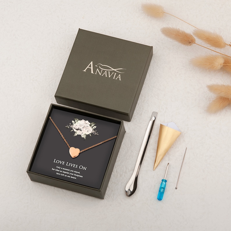 Puede incluir: Un collar con un colgante de coraz&oacute;n dorado en una caja verde con un dise&ntilde;o floral blanco. El collar est&aacute; sobre una tarjeta negra con el texto "Love Lives On. Only a moment you stayed, but what an impact your footprints have left on our hearts."