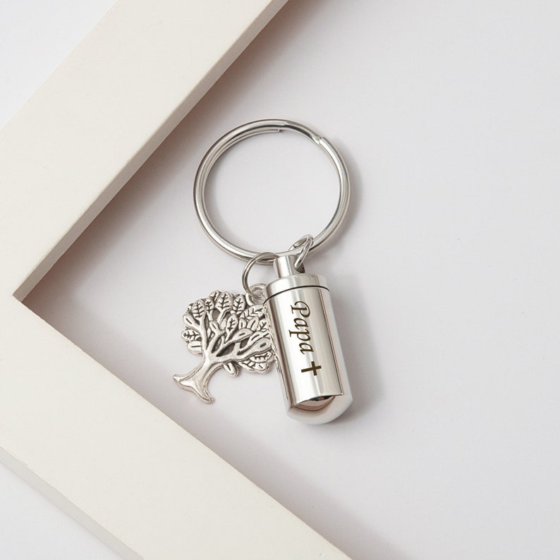 May include: A silver keychain with a small tree charm and a cylindrical cremation urn. The urn has the words 'Papa +' engraved on it.