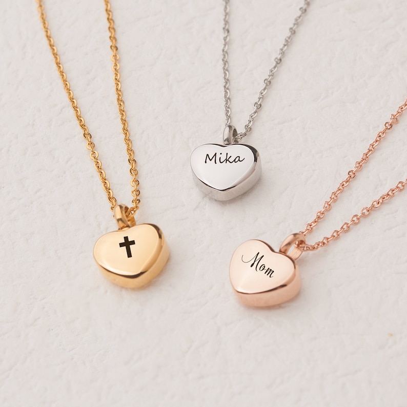 May include: Three gold heart-shaped necklaces with different engravings. One necklace has a cross, another says "Mom", and the third says "Mika".