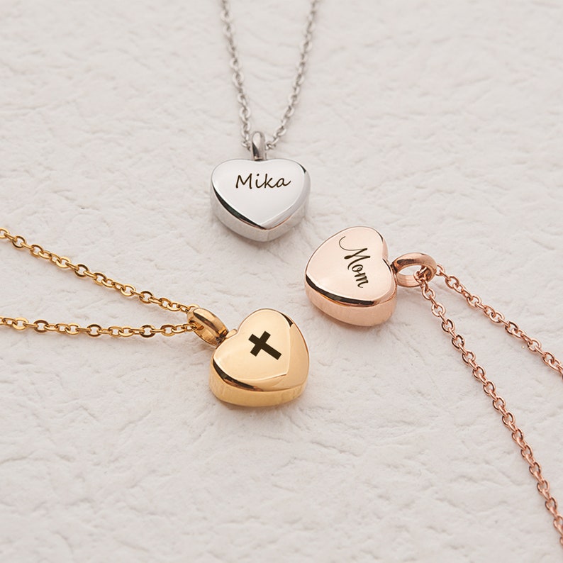 May include: Three heart-shaped cremation jewelry pendants on chains. The pendants are silver, rose gold, and gold. The silver pendant is engraved with the name "Mika", the rose gold pendant is engraved with the word "Mom", and the gold pendant has a cross engraved on it.