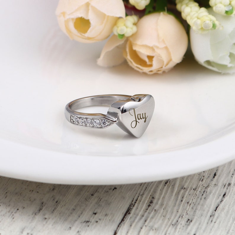 May include: A silver heart-shaped ring with a small row of clear cubic zirconia stones on the band. The heart is engraved with the name "Jay".