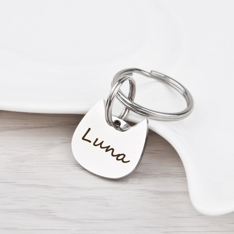 Puede incluir: Una placa de identificaci&oacute;n para mascotas de metal plateado con forma de cabeza de gato y el nombre Luna grabado en una fuente cursiva oscura. La placa est&aacute; unida a una anilla de metal, creando un accesorio funcional para un collar.