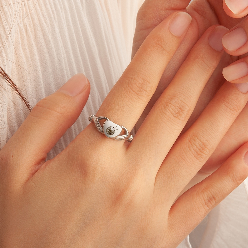 May include: A silver ring with a white heart-shaped setting featuring a dandelion design.