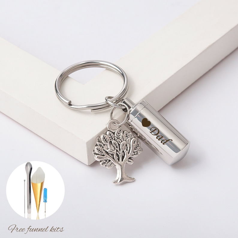 May include: Silver keychain with a small tree charm and a cylindrical cremation urn engraved with "Dad" and a heart. The urn is attached to the keychain with a ring. A small funnel kit is included.