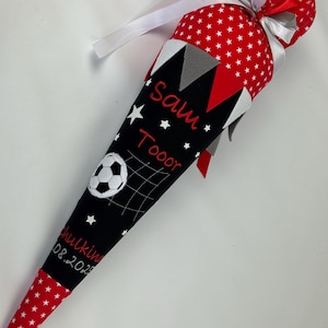 Può includere: Un sacco scolastico a forma di cono in tessuto rosso e nero con un disegno di pallone da calcio e il nome "Sam Tooor" ricamato sopra. La borsa ha un motivo a stelle rosse sulla parte superiore e inferiore, con un fiocco di nastro bianco.