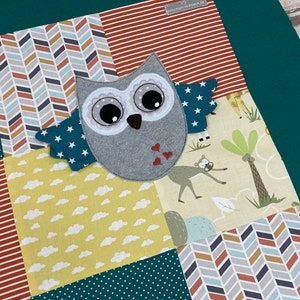 Könnte beinhalten: Eine Patchwork-Babydecke mit einer grauen Eulen-Applikation in der Mitte. Die Decke hat verschiedene Muster, darunter Chevron, Punkte und Wolken. Die Eule hat große, schwarze Augen und einen kleinen, orangefarbenen Schnabel.