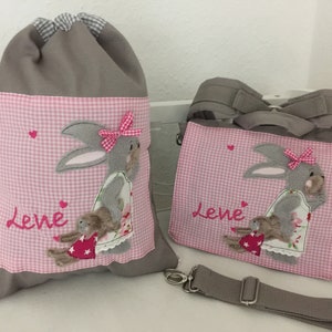Op de afbeelding: Twee grijze stoffen tassen met roze en witte ruitjesstof accenten. Beide tassen hebben een grijs konijn met een roze strik en de naam "Lene" geborduurd op de voorkant.