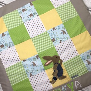Peut inclure: Une couverture bébé en patchwork avec une applique de lapin brun et le nom "FREYA" brodé en lettres jaunes. La couverture est faite de différents tissus dans des tons de vert, jaune, bleu et blanc.