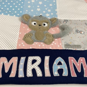 Könnte beinhalten: Eine blaue und rosafarbene Patchwork-Babydecke mit einem grauen Affen-Applikationen und dem Namen "MIRIAM" in rosa und blauen Buchstaben gestickt.