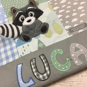 Könnte beinhalten: Eine graue und weiße Patchwork-Babydecke mit einem Waschbär-Applikationen und dem Namen "LUCA" in Blau, Grün und Grau gestickt.