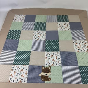 Könnte beinhalten: Eine Patchwork-Babydecke mit braunem Rand, bestehend aus grünen, blauen und beigefarbenen Quadraten. Die Quadrate weisen Muster wie Punkte, Streifen und einen Waldtierdruck auf. Auf der Decke ist eine gestickte Fuchsapplikation aufgenäht.