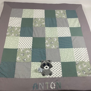 Könnte beinhalten: Eine graue und grüne Patchwork-Babydecke mit einem Waschbär-Plüschtier und dem Namen Anton, der unten gestickt ist.
