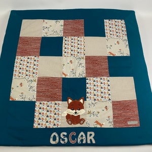 Może przedstawiać: Turkusowy koc dla niemowląt z patchworkowym wzorem w różnych wzorach w kolorze brązowym, beżowym i białym. Koc ma aplikację lisa i imię "OSCAR" wyszyte na biało.