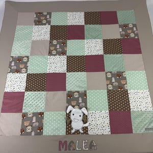 Op de afbeelding: Een beige en bruine patchwork babydeken met een groen, wit en bruin patroon. Een wit pluche hondenspeelgoed bevindt zich in het midden van de deken. De naam "MALEA" is in roze op de deken geborduurd.