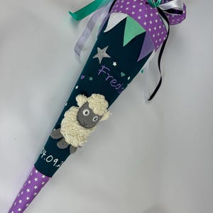 Puede incluir: Un cono escolar de color verde azulado oscuro con un patrón de estrellas moradas en la parte inferior y superior. El cono presenta una oveja de fieltro, el nombre "Freda" y banderines decorativos. Cintas en verde azulado, blanco y morado adornan la parte superior.