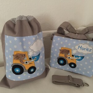 Op de afbeelding: Twee grijze stoffen tassen met blauw-witte sterrenpatronen. De tassen hebben een geborduurd beeld van een geel bouwvoertuig met een bruine beer die rijdt. Een tas heeft de naam "Moritz" geborduurd.