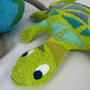 Pode incluir: Um brinquedo de pelúcia verde e azul em forma de lagarto ou dinossauro. O brinquedo tem uma aparência texturizada e difusa com detalhes triangulares azuis e turquesa. Os olhos são brancos com pupilas azuis escuras. Uma Terra de pelúcia azul e verde é parcialmente visível.