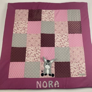 Op de afbeelding: Een roze en grijze patchwork speelmat met een grijze ezel-applique en de naam "NORA" geborduurd in roze.