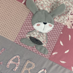 Op de afbeelding: Een grijs en roze patchwork kussen met een grijze konijnen-applicatie en de naam "LARA" geborduurd in roze op de voorkant.