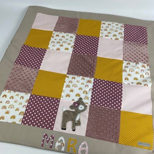 Può includere: Un tappetino da gioco per bambini beige con un design a patchwork con tessuti rosa, gialli e bianchi. Il tappetino ha un'applicazione a forma di cervo e il nome "MARA" ricamato.