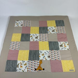 Peut inclure: Une couverture patchwork pour bébé avec des carrés de différentes couleurs et motifs : jaune, rose, gris et blanc. Bordure beige et application centrale d'une girafe. Thème safari pour cette couverture.