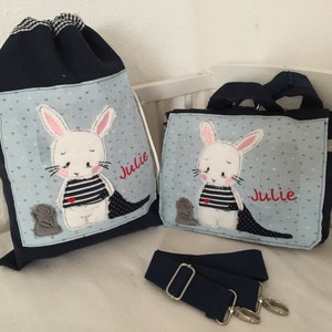 Op de afbeelding: Een set van twee blauwe canvas tassen met een wit konijn en de naam "Julie" geborduurd op elke tas. De kleinere tas heeft een schouderband.