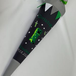 Könnte beinhalten: Eine dunkelgraue, kegelförmige Schultüte mit Minecraft-Motiv. Die Tüte hat grüne und weiße Akzente, ein grünes Creeper-Design und den Namen "Christoph" in Grün gestickt. Das Datum "09.08.2023" ist ebenfalls sichtbar.