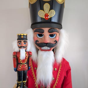 May include: A large nutcracker figure wearing a black hat with gold trim and a red jacket with gold accents, featuring a white beard and moustache. The figure holds a smaller, matching nutcracker model in its hands, both displaying painted facial features.