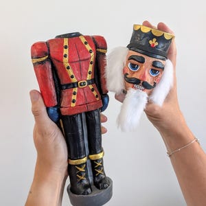 May include: A two-piece nutcracker figurine featuring a red jacket with gold trim and black trousers. The head is detached, showcasing a black hat with a gold emblem, blue eyes, a black moustache, and white synthetic hair and beard.