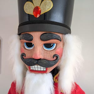 May include: A nutcracker figure featuring a tall black hat with gold trim and a red gem, a red jacket with gold accents, and a white faux fur beard and hair. The face has blue eyes, pink cheeks, and a black moustache over a white toothy mouth.