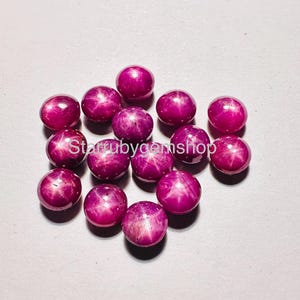Puede incluir: Un conjunto de catorce rubíes estrella redondos, tallados en cabujón, de un tono magenta rosado intenso. Cada piedra preciosa muestra un efecto de estrella de seis puntas en su superficie lisa y pulida. El texto "Starrubygemshop" es visible en el centro.