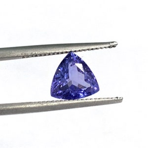 May include: A triangular purple gemstone with facets, held by tweezers against a white background.