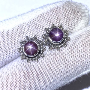 Könnte beinhalten: Ein Paar silberne Ohrringe mit einem sternförmigen Rubin-Edelstein in jedem. Die Ohrringe haben ein Sternburst-Design um den Rubin.