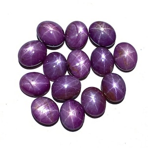 Könnte beinhalten: Eine Sammlung von zwölf violetten Sternrubin-Cabochons. Die Steine sind oval geformt und haben ein sternförmiges Muster in der Mitte.