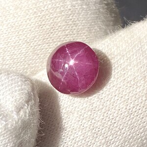 Könnte beinhalten: Ein runder, rosafarbener Sternrubin-Edelstein mit einem sechszackigen Stern in der Mitte.