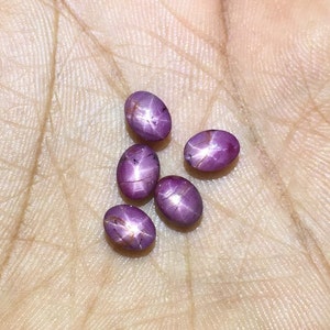 Könnte beinhalten: Sechs ovale, violette Sternrubin-Edelsteine mit einem sternförmigen Muster in der Mitte.