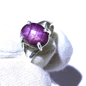 Könnte beinhalten: Ein Silberring mit einem violetten Sternrubin-Edelstein. Der Rubin ist oval geformt und hat ein sternförmiges Muster.