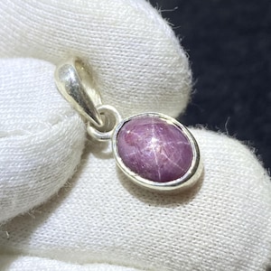 Könnte beinhalten: Ein silberner Anhänger mit einem ovalen, sternförmigen Rubin-Edelstein. Der Rubin hat eine tiefe rosa Farbe mit einem sichtbaren Sterneffekt. Der Anhänger hat ein schlichtes, elegantes Design, das für den Alltag geeignet ist.
