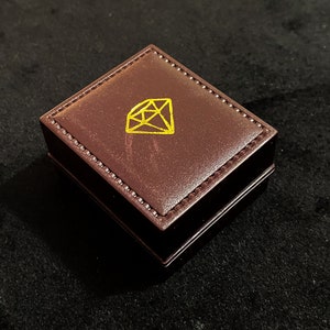 Op de afbeelding: Een bruine leren ringdoos met een gouden diamantsymbool op de bovenkant.