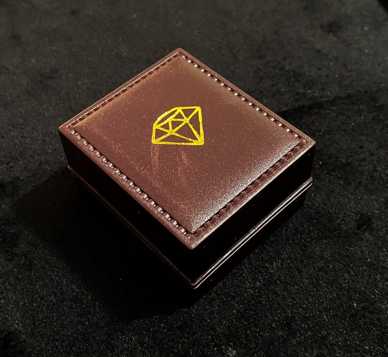 Op de afbeelding: Een bruine leren ringdoos met een gouden diamantsymbool op de bovenkant.