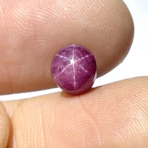 Könnte beinhalten: Ein polierter, ovaler Rubin-Edelstein. Der Edelstein hat eine tief purpurrote Farbe mit einem sechsstrahligen Sterneffekt auf seiner Oberfläche. Das Sternmuster ist weiß und hebt sich von der satten Farbe des Rubins ab. Der Edelstein wird zwischen Fingern gehalten.