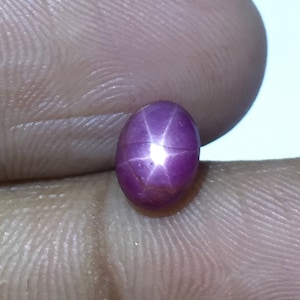 Könnte beinhalten: Ein sternförmiger Rubin-Edelstein mit einem tiefen Rosaton. Der Edelstein ist oval geformt und hat eine polierte Oberfläche.