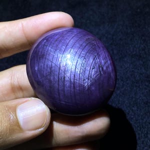 Può includere: Una gemma di zaffiro stellato viola, lucida e di forma ovale, caratterizzata da un distinto motivo a stella a sei punte sulla superficie. La pietra viola intenso presenta un taglio a cabochon liscio con inclusioni fibrose interne.