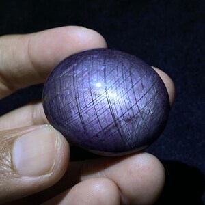 Könnte beinhalten: Ein polierter, oval geformter Cabochon-Edelstein mit einem tiefvioletten Farbton. Die Oberfläche zeigt ein ausgeprägtes, sich überschneidendes Gitter aus feinen, dunklen Linien, die ein schraffiertes Muster auf der glatten, abgerundeten Steinfläche bilden.
