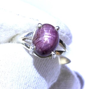 Könnte beinhalten: Ein silberner Ring mit einem violetten Sternrubin-Edelstein. Der Rubin ist oval geformt und hat ein sternförmiges Muster.