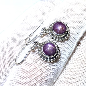 Könnte beinhalten: Ein Paar silberne Ohrringe mit jeweils einem violetten Sternrubin-Edelstein. Die Ohrringe haben einen perlenbesetzten Rand um den Edelstein.