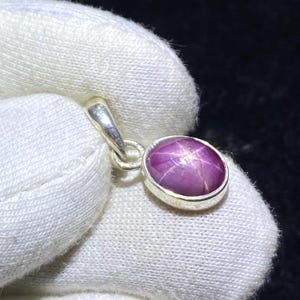 May include: A pendant with a polished, oval purplish-red star ruby gemstone. The gemstone is set in a silver-coloured bezel and displays a six-ray star pattern. The pendant has a silver loop for a chain.