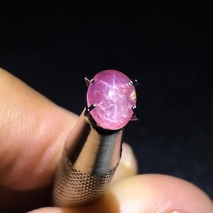 Könnte beinhalten: Ein leuchtend rosa, ovaler Sternsaphir-Edelstein in einer Metall-Krappenfassung. Der im Cabochon-Schliff gehaltene Stein zeigt einen ausgeprägten sechsstrahligen Sterneffekt auf seiner Oberfläche, der das Licht vor einem dunklen Hintergrund reflektiert.